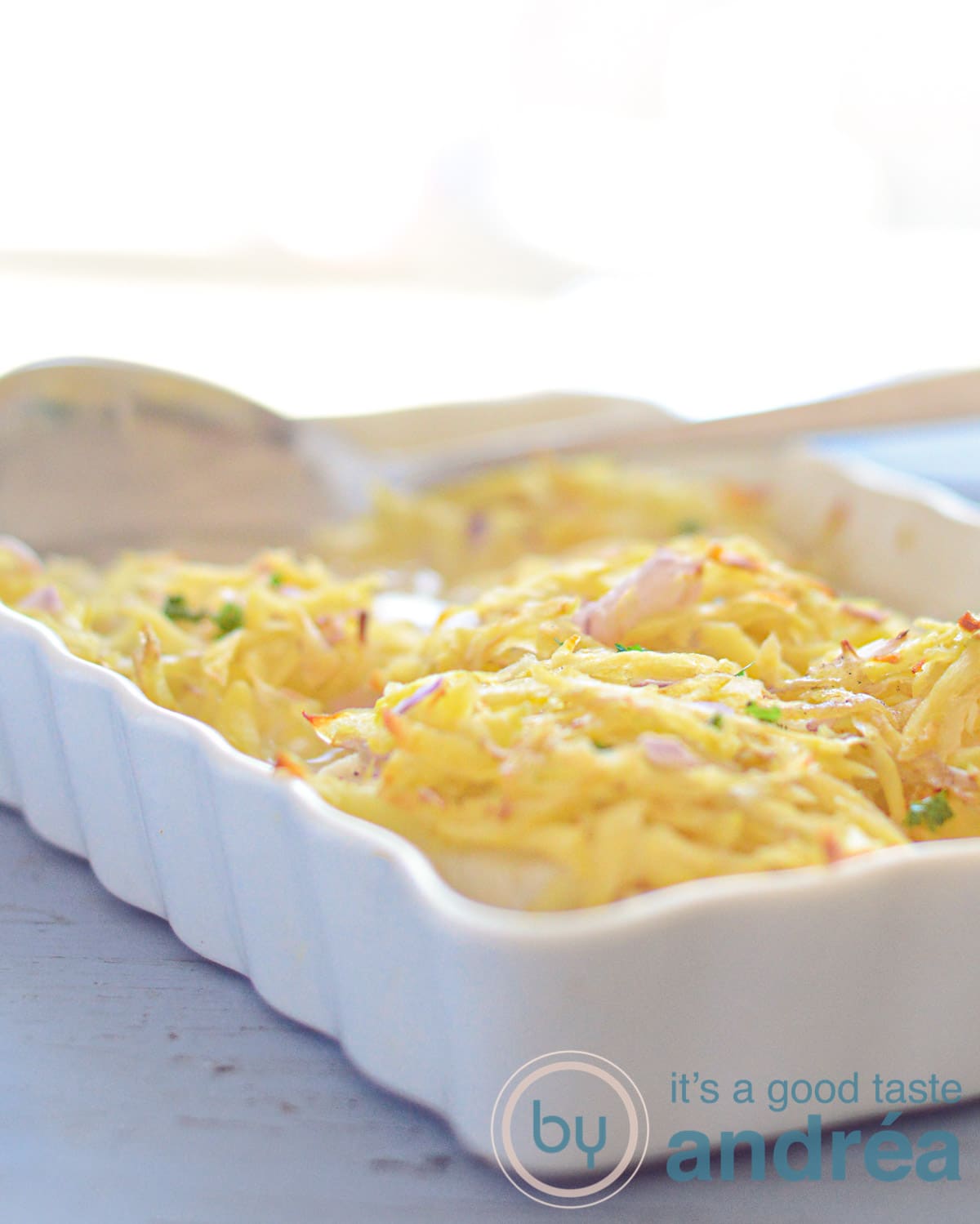 Een witte ovenschaal op een blauwe ondergrond met kabeljauwfilet met rösti topping en een opscheplepel