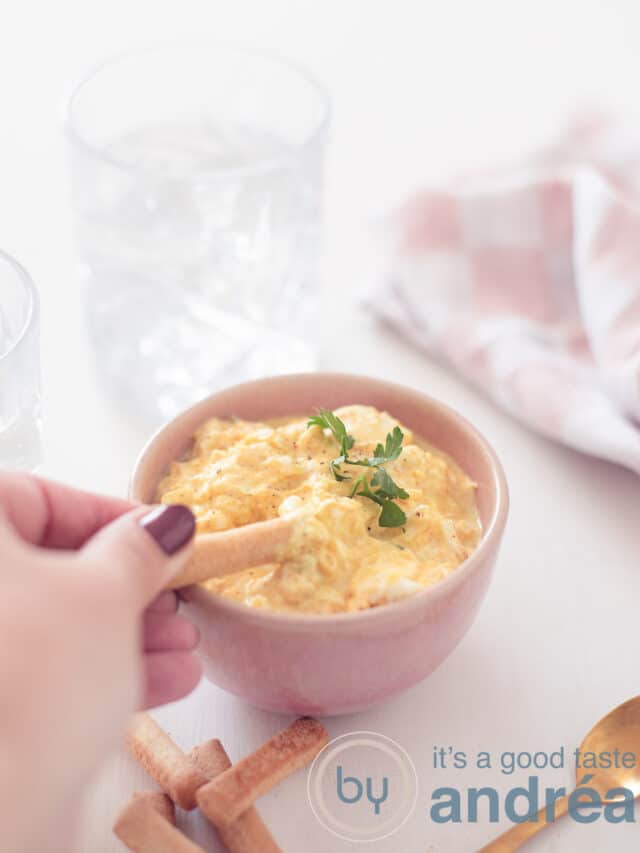 a pink bowl with egg salad, no may, no mustard. A bread stick dipping in it. A white background.