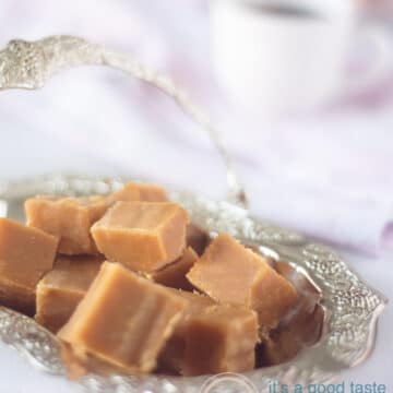 Een zilveren schaaltje met blokjes caramel fudge. Een kopje koffie op de achtergrond