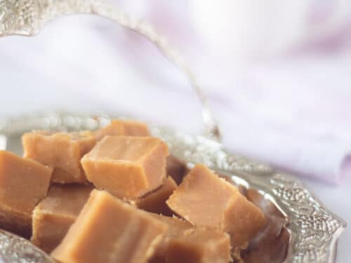 Een zilveren schaaltje met blokjes caramel fudge. Een kopje koffie op de achtergrond