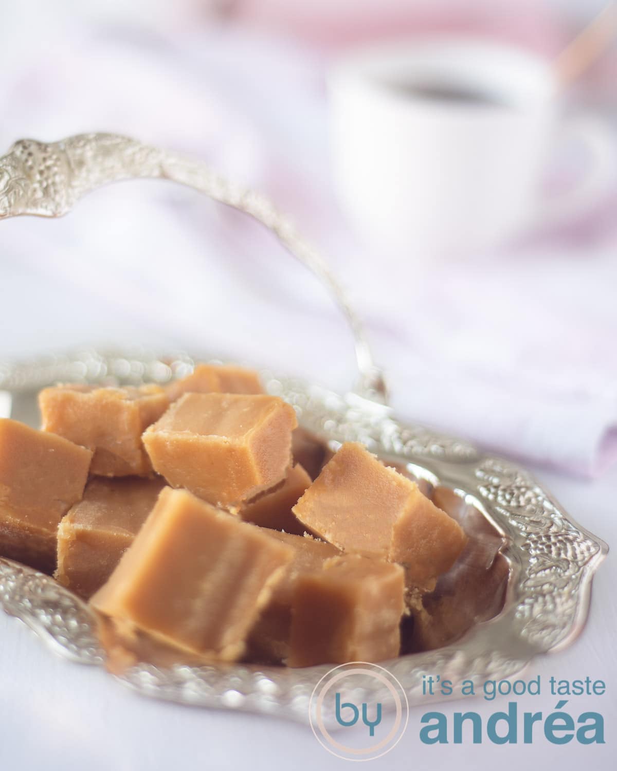 Een zilveren schaaltje met blokjes caramel fudge. Een kopje koffie op de achtergrond