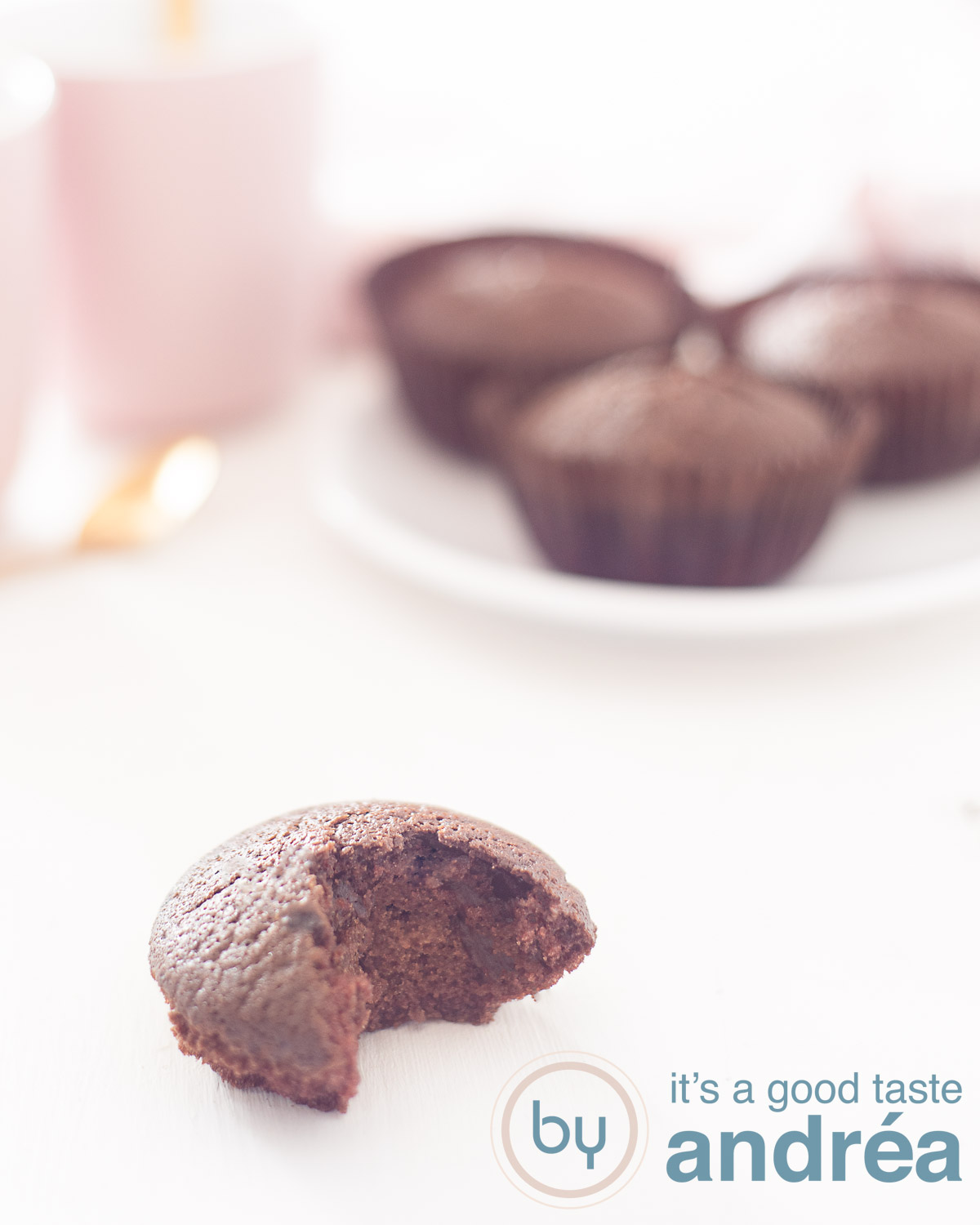 dairy free chocolate cupcake in front with a bite taking out. In the background a white plate with three chocolate cupcakes and a pink mug.