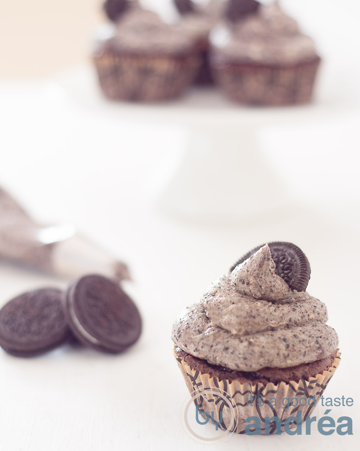 Een Oreo cupcake op een witte ondergrond. Linksachter enkele oreo's en een spuitzak. Helemaal achterin een taartstandaard met Oreo chocolade cupcakes