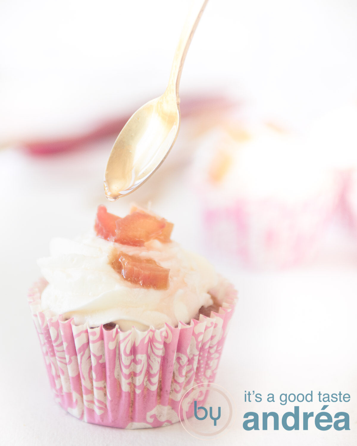 Een roze wite cupcake vorm gevuld met een rabarber muffin met een slagroom topping met rabarber. Een lepel sprenkelt wat siroop op de rabarber