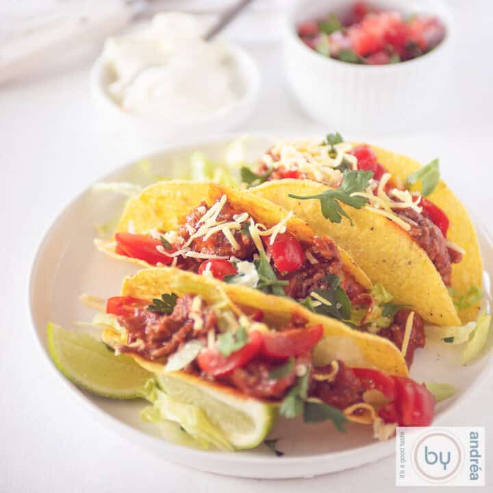Een vierkante foto met drie taco's gevuld met gehaktsaus, tomaat en kaas. Een salsa en creme fraiche in de achtergrond