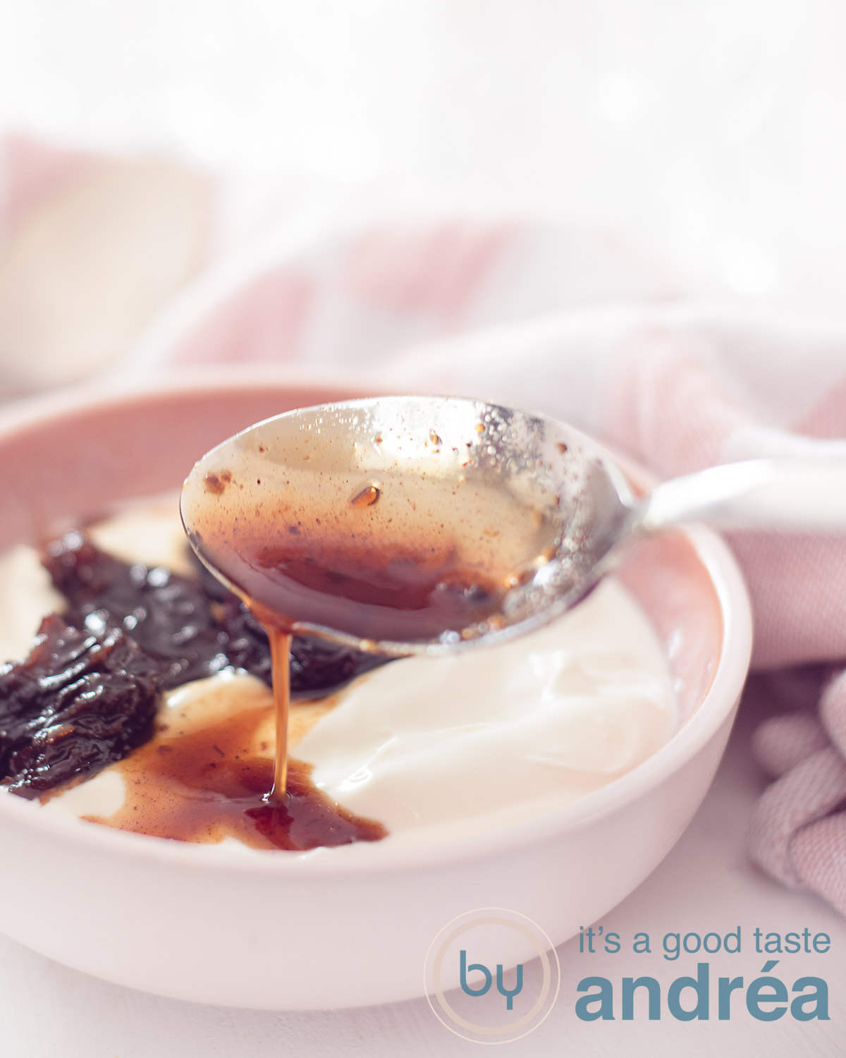 Griekse yoghurt met pruimen in een roze schaaltje. Een lepel schenkt wat saus bij dit toetje. Een roze witte doek op de achtergrond.