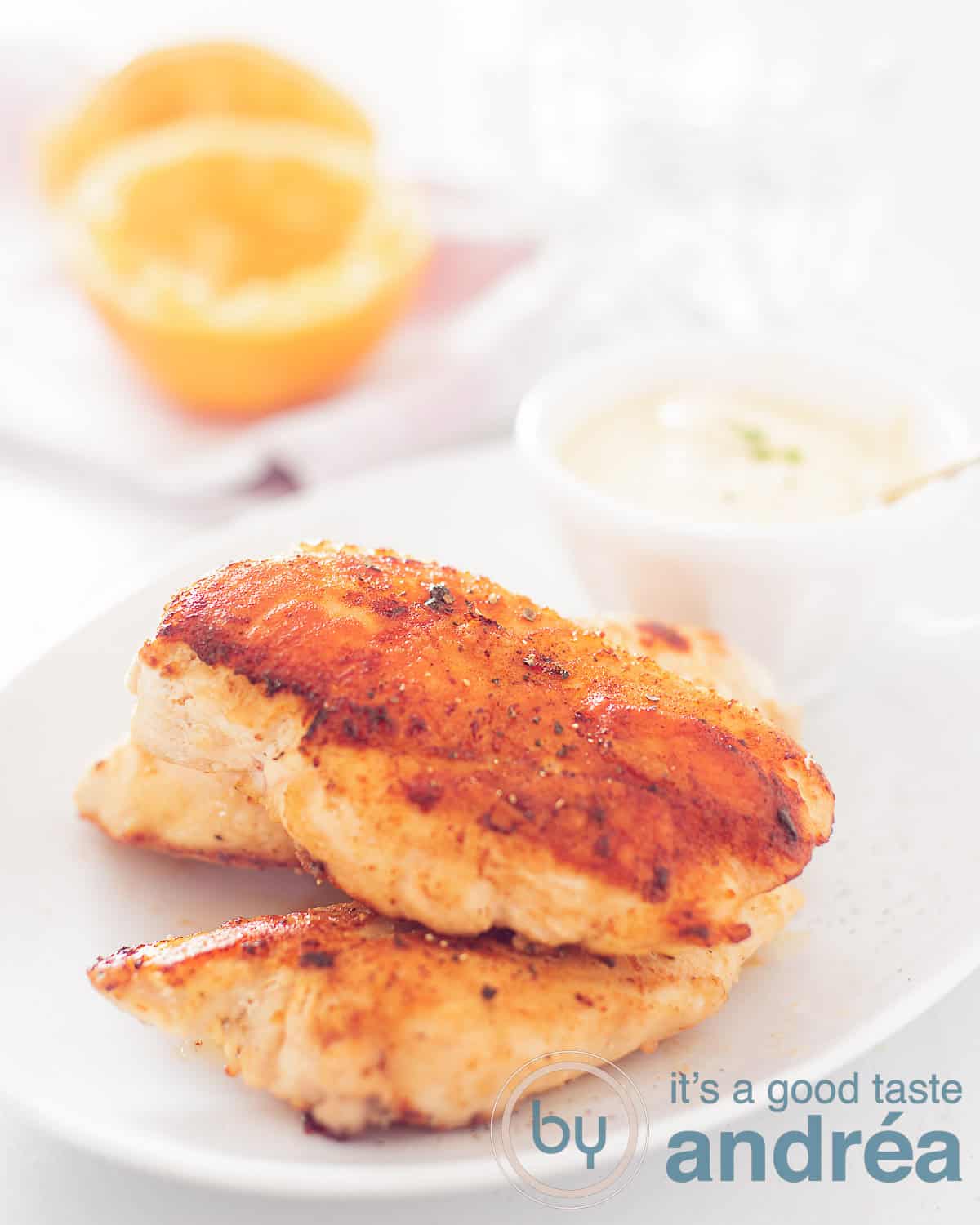 Een hoogtefoto met een wit bord met drie kipfilets en een kommetje sinaasappel tijm saus. In de achtergrond twee uitgeperste sinaasappelhelften. Alles op een witte ondergrond