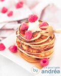 Pancakes met frambozen en crème fraîche op een wit bord. Er omheen meer frambozen. In de achtergrond een wit roze theedoek.
