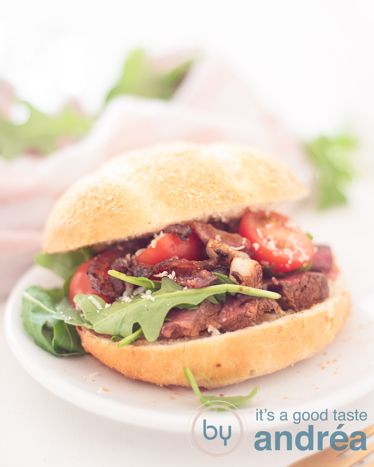 Sandwich with grilled steak and caramelized onions on a white plate. In the backgroudn a pink/white cloth and some arugula.