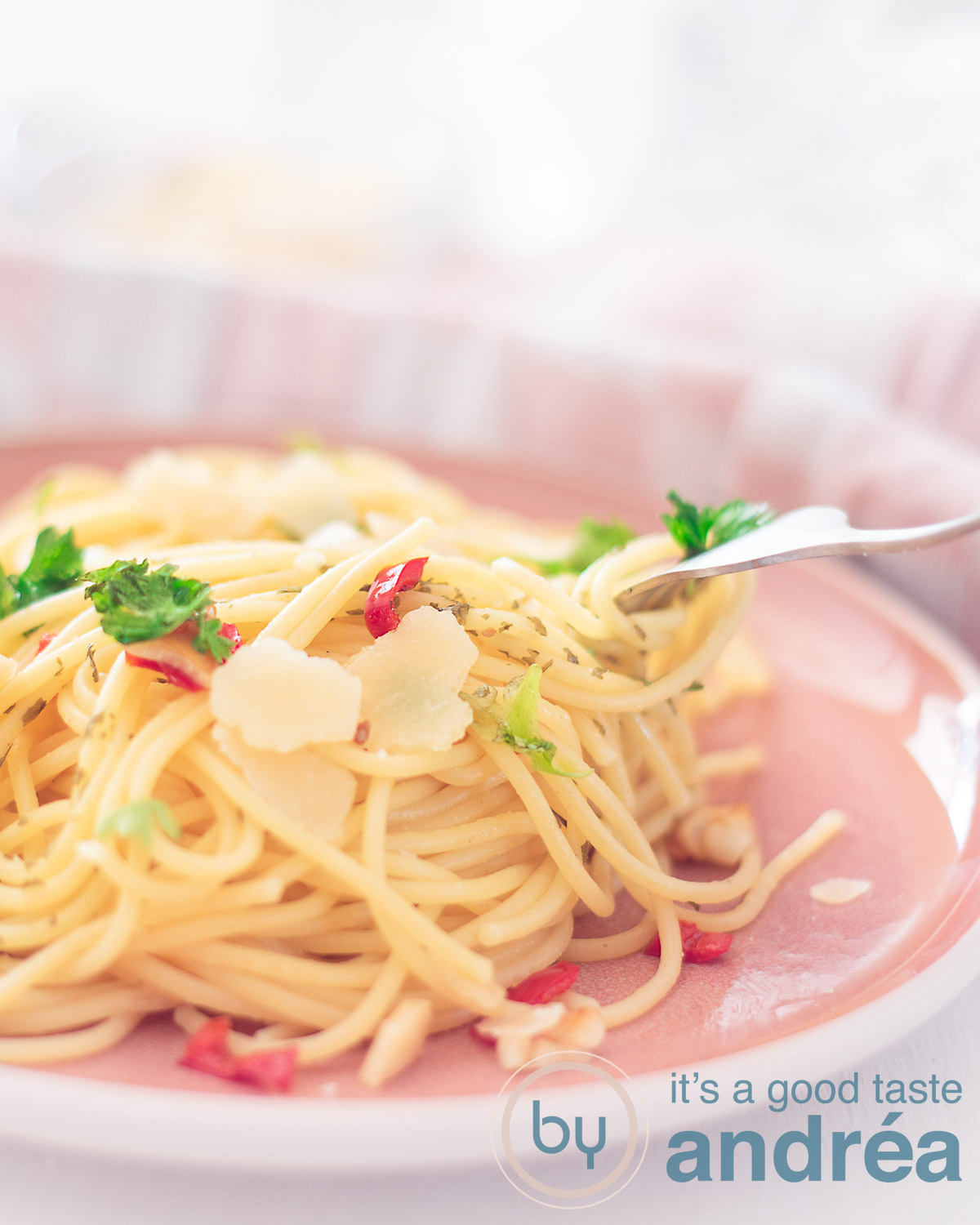 Een deel van een roze bord met een berg spaghetti met pepers, peterselie, parmezaanse kaas, knoflook en olijfolie. Een vork steekt in de spaghetti. Een witte ondergrond