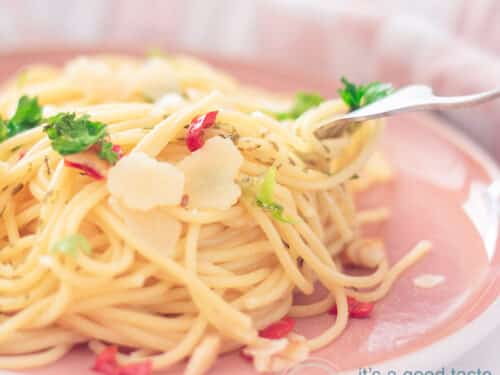 Een vierkante foto met een deel van een roze bord, een berg spaghetti op smaak gebracht met peper, olijfolie en knoflook, peterselie en geschaarde parmezaanse kaas