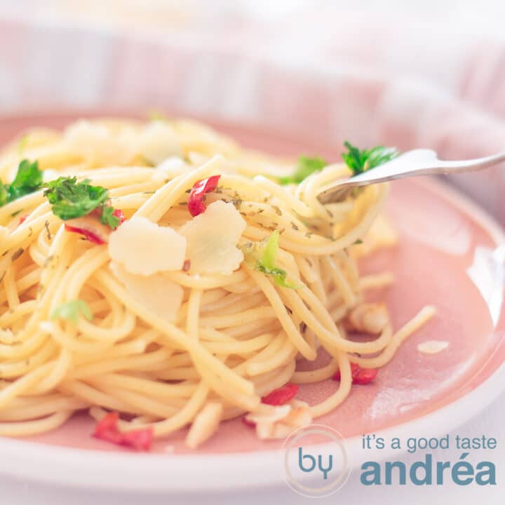 Een vierkante foto met een deel van een roze bord, een berg spaghetti op smaak gebracht met peper, olijfolie en knoflook, peterselie en geschaarde parmezaanse kaas
