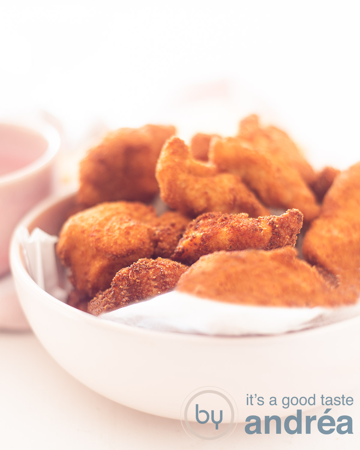 Een roze schaaltje bekleedt met bakpaier en gevuld met kipnuggets. Achterin een witte achtergrond.