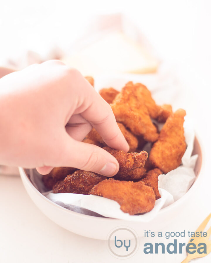 Een hand pakt een kipnugget uit een roze schaal met kipnuggets een witte achtergrond.