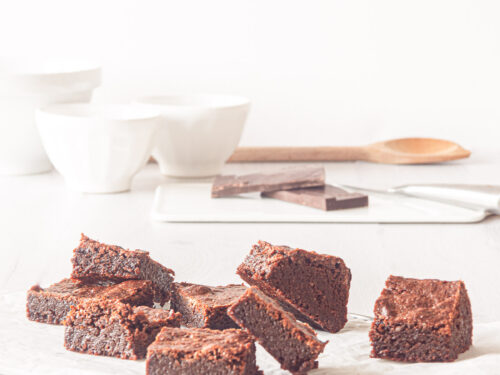 Op een wit bakpapier liggen browneis verspreid. Op de achtergrond een wit kommetje, stukjes chocola en een houten lepel