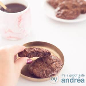 Een vierkante foto met een Brownie cookie, een hand pakt hem van de schaal af. Links achterin een roze mok met koffie. Rechts bovenin een wit bord met een aantal brookies. Een witte ondergrond