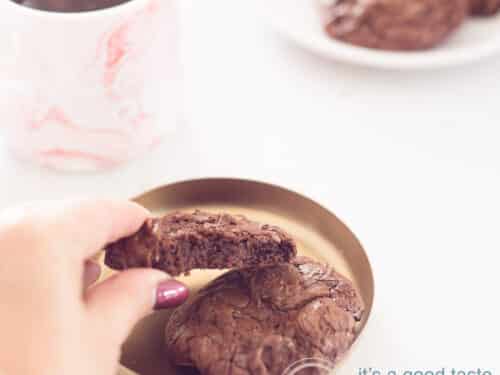 Een vierkante foto met een Brownie cookie, een hand pakt hem van de schaal af. Links achterin een roze mok met koffie. Rechts bovenin een wit bord met een aantal brookies. Een witte ondergrond