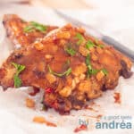 Een vierkante foto met twee stukken sparerib die op elkaar liggen. Ze zijn besprenkelt met zoete barbecuesaus en peterselie