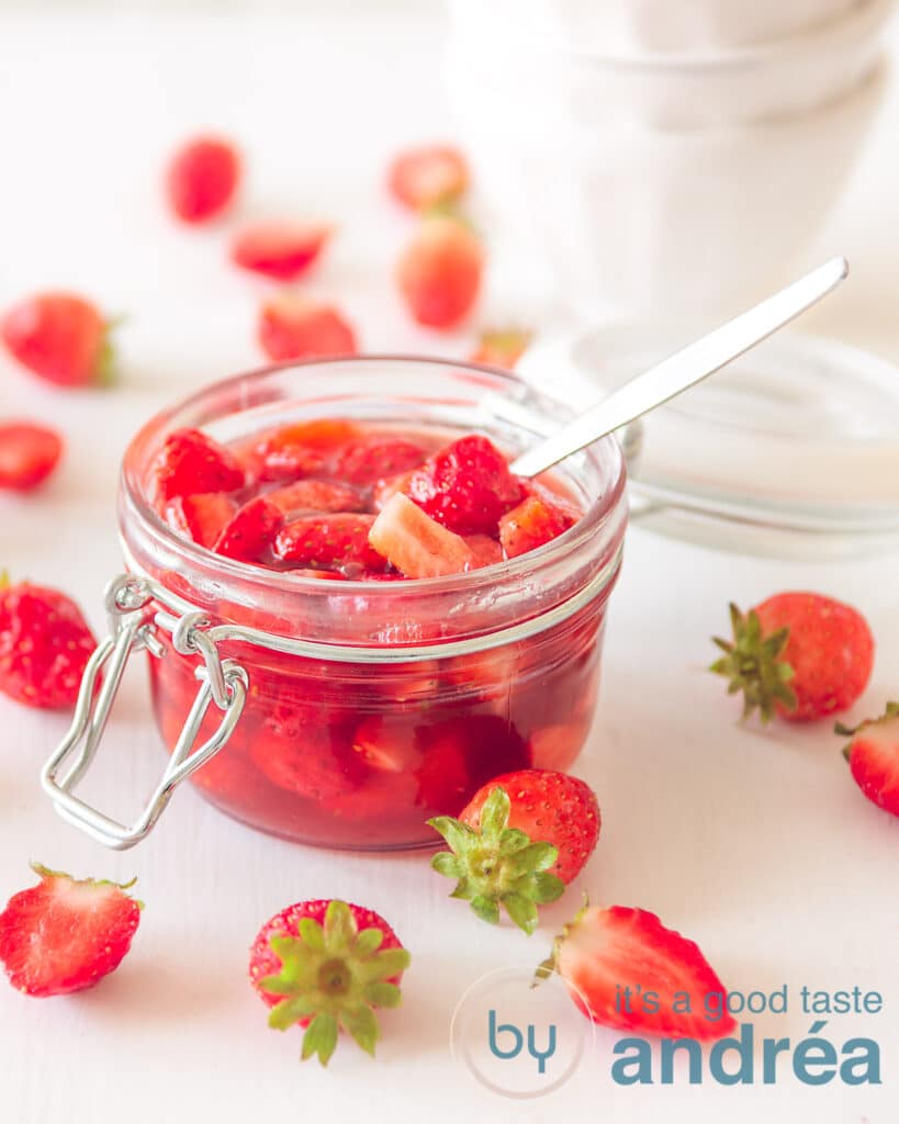 een glazen pot met zelfgemaakte aardbeiensaus met een lepel. Wat witte schaaltjes in de achtergrond.