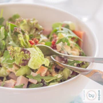 Een vierkante foto met een witte kom met groene salade, gerookte kip en zongedroogde tomaten.