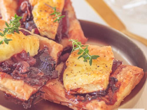 Een vierkante foto met bladerdeeghapjes met rode ui, brie en spekjes