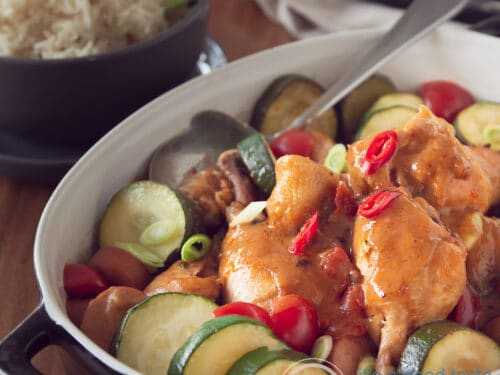Een vierkante foto met een zwarte ovenschaal met gumbo, kip, chorizo en courgette. Rijst in de achtergrond