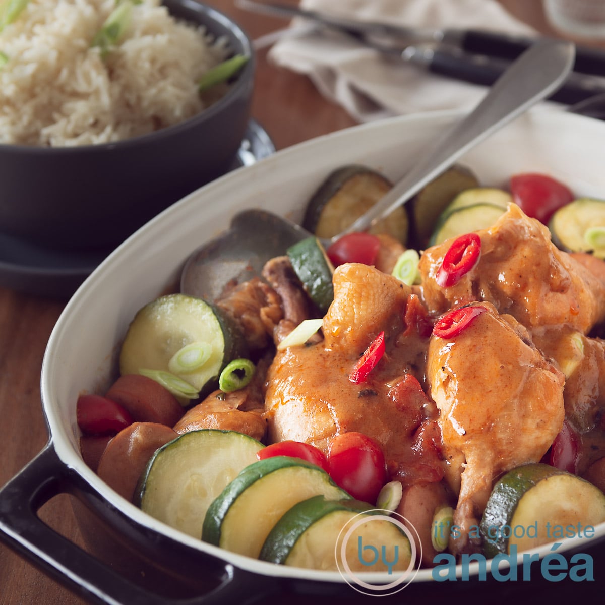 Een vierkante foto met een zwarte ovenschaal met gumbo, kip, chorizo en courgette. Rijst in de achtergrond