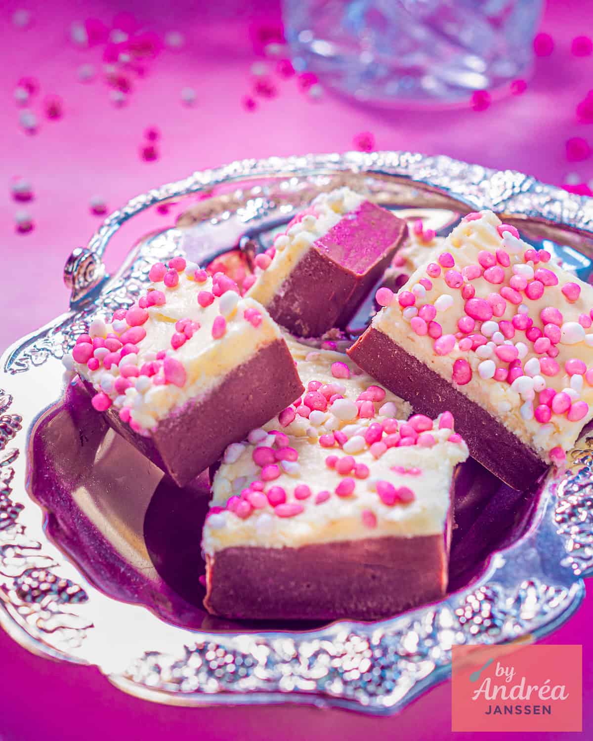 Een roze achtergrond, een zilveren schaal met red velvet fudge met frosting en muisjes