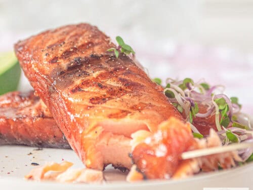 Een vierkante foto met een wit bord met twee stukken gegrilde zalm die gepekeld is.