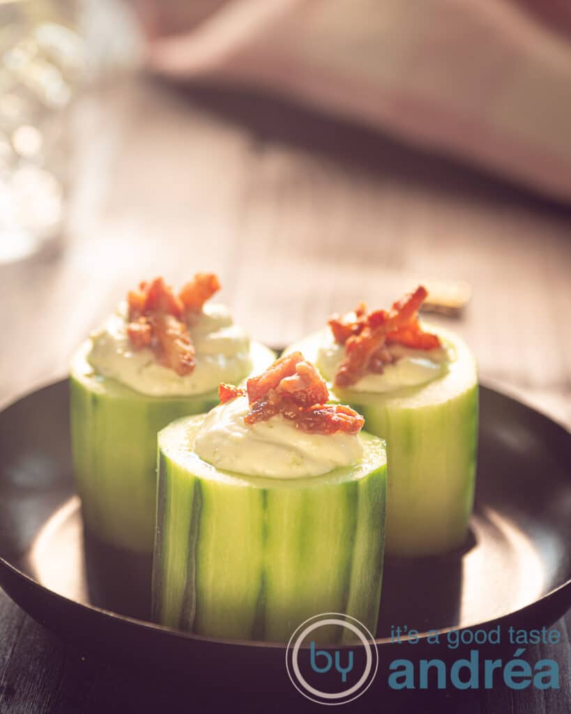 Three cucumber appetizers with leek dip and topped with bacon on a black plate.