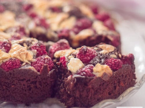 Een vierkante foto met een deel van een brownie waar een punt uit is gesneden. Een topping van witte chocola en frambozen