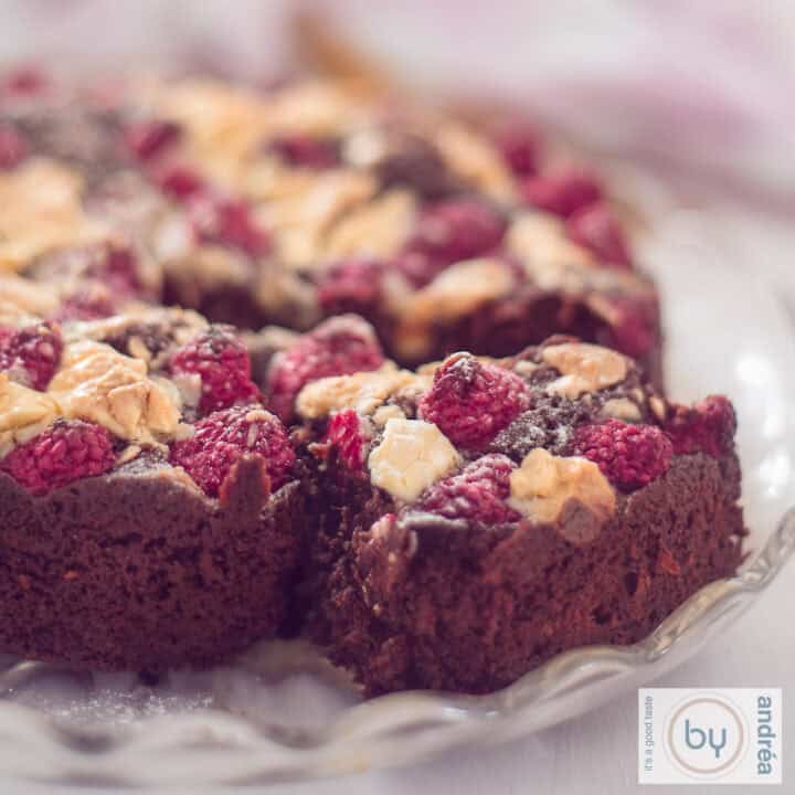 Een vierkante foto met een deel van een brownie waar een punt uit is gesneden. Een topping van witte chocola en frambozen