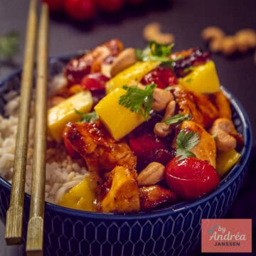 Een vierkante foto van een blauwe schaal met rijst, topping van mango, kip in honing ketjapsaus, cashews en tomaten.