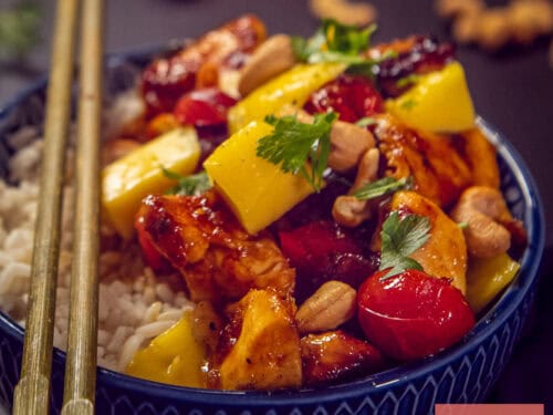 Een vierkante foto van een blauwe schaal met rijst, topping van mango, kip in honing ketjapsaus, cashews en tomaten.