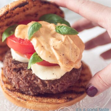 een hand die het kapje op het geroosterde broodje van de burger wil leggen met tomaat mozzarella en meer