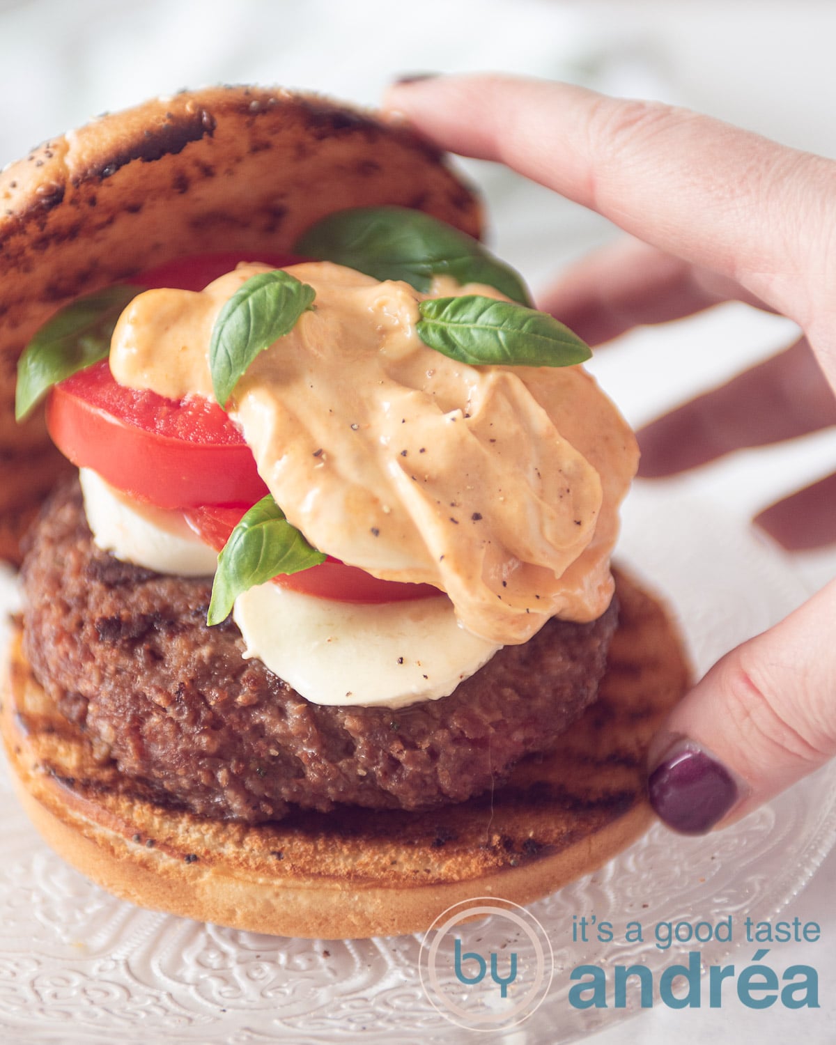 een hand die het kapje op het geroosterde broodje van de burger wil leggen met tomaat mozzarella en meer