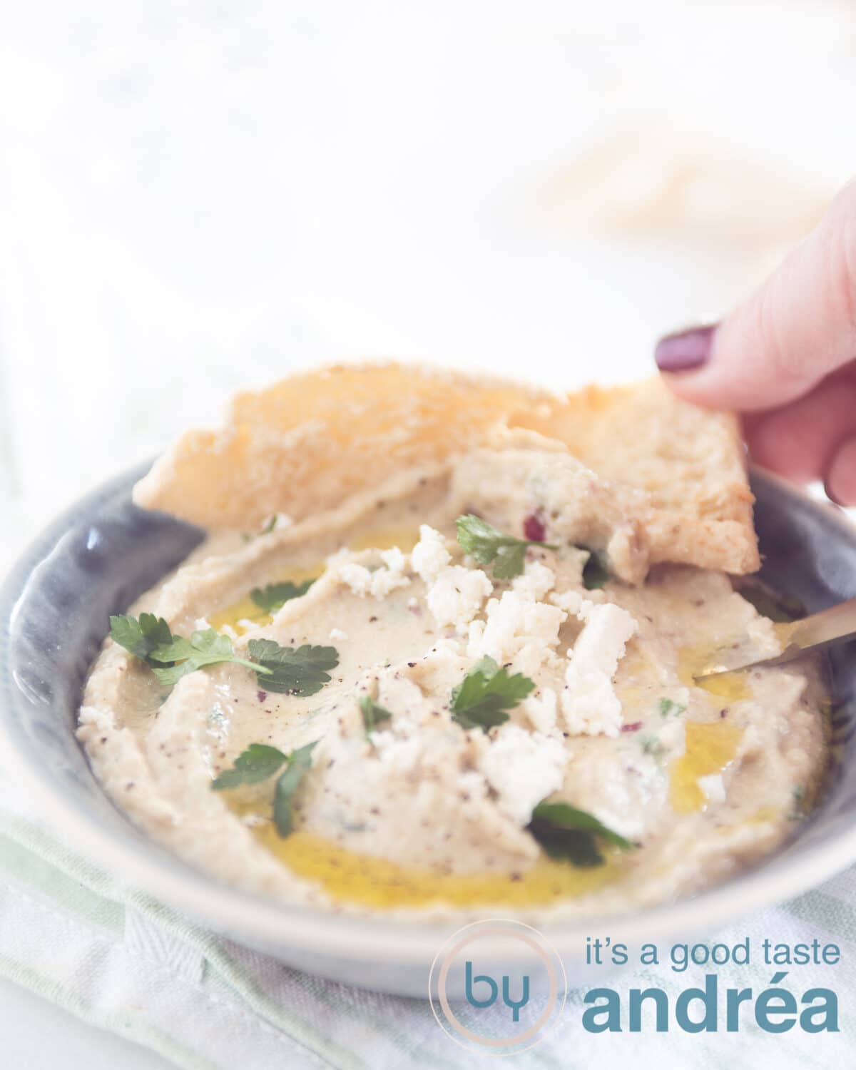 Een grijs schaaltje gevuld met Griekse aubergine dip, pita puntjes en een hand die een pita puntje pakt