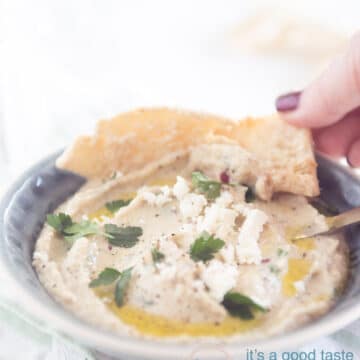 Een grijs schaaltje gevuld met Griekse aubergine dip, pita puntjes en een hand die een pita puntje pakt