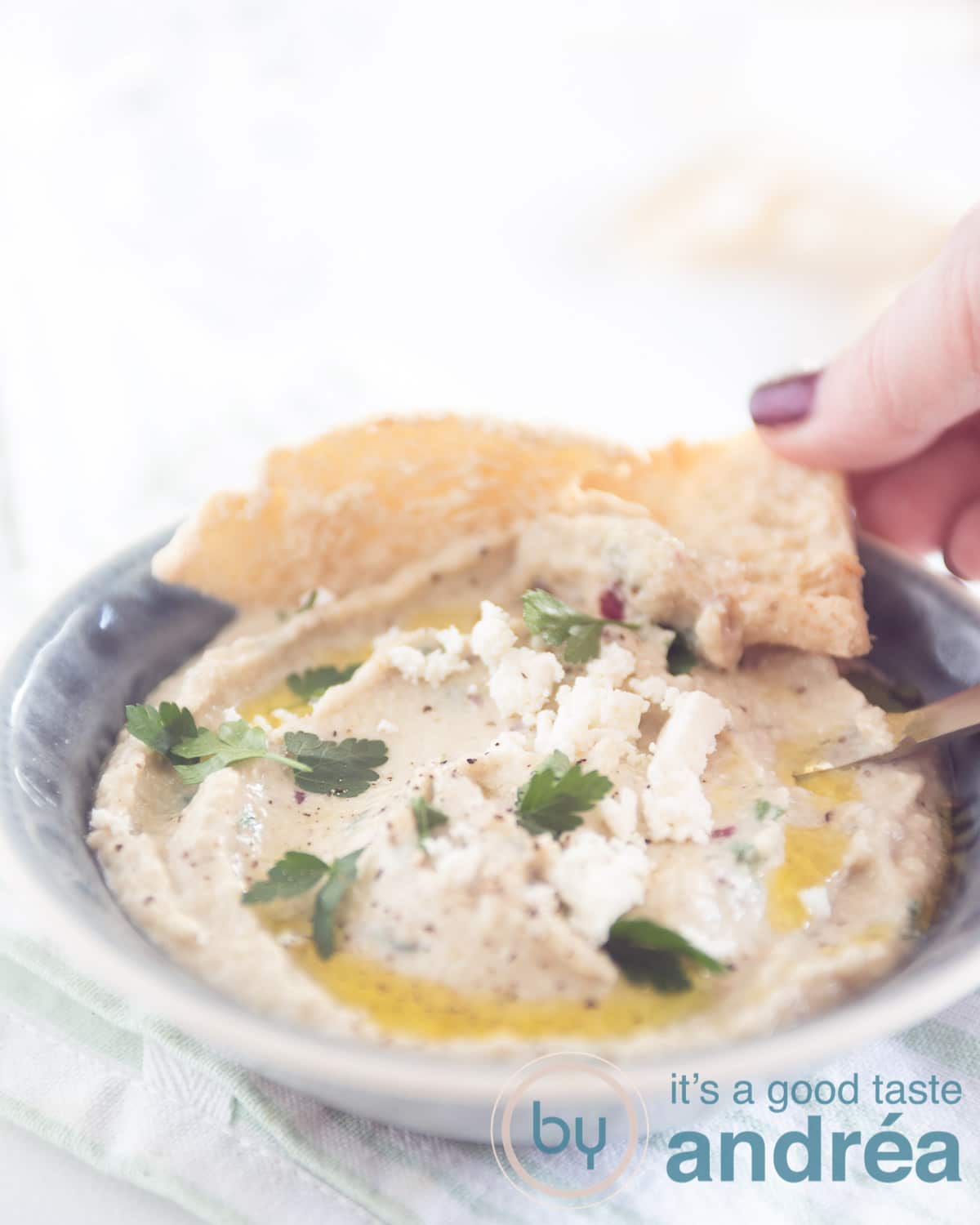 Een grijs schaaltje gevuld met Griekse aubergine dip, pita puntjes en een hand die een pita puntje pakt