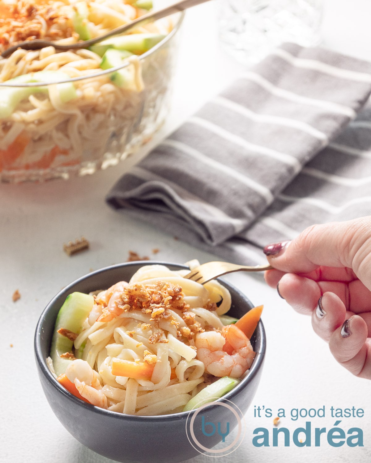 a hand taking a bit of the noodle salad in a great bowl. A big bowl filled with salad and a grey towel are in the back