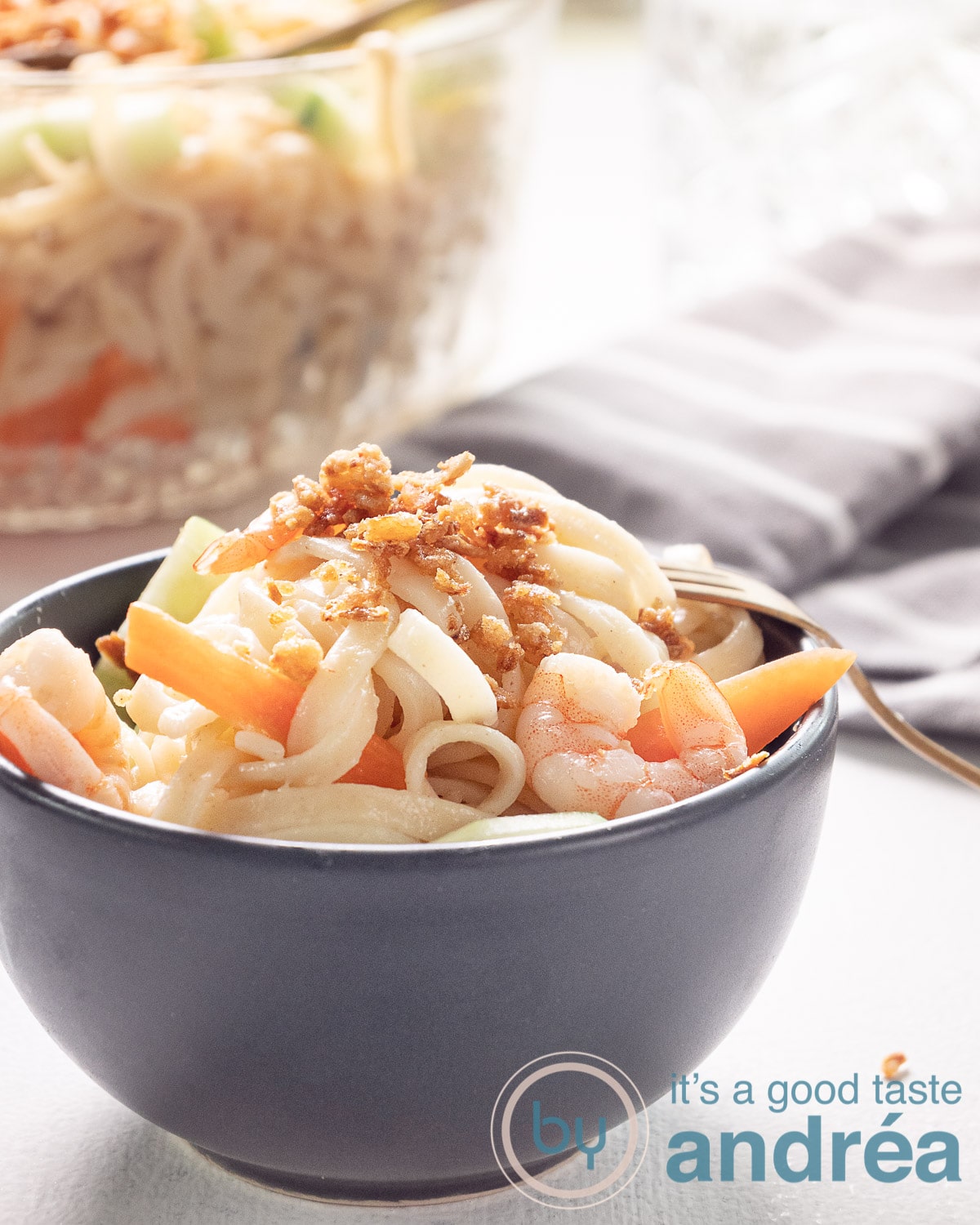 A grey bowl filled with Indonesian noodle salad with cucumber, carrot and shrimp. A glass bowl at the back. A grey towel on the right