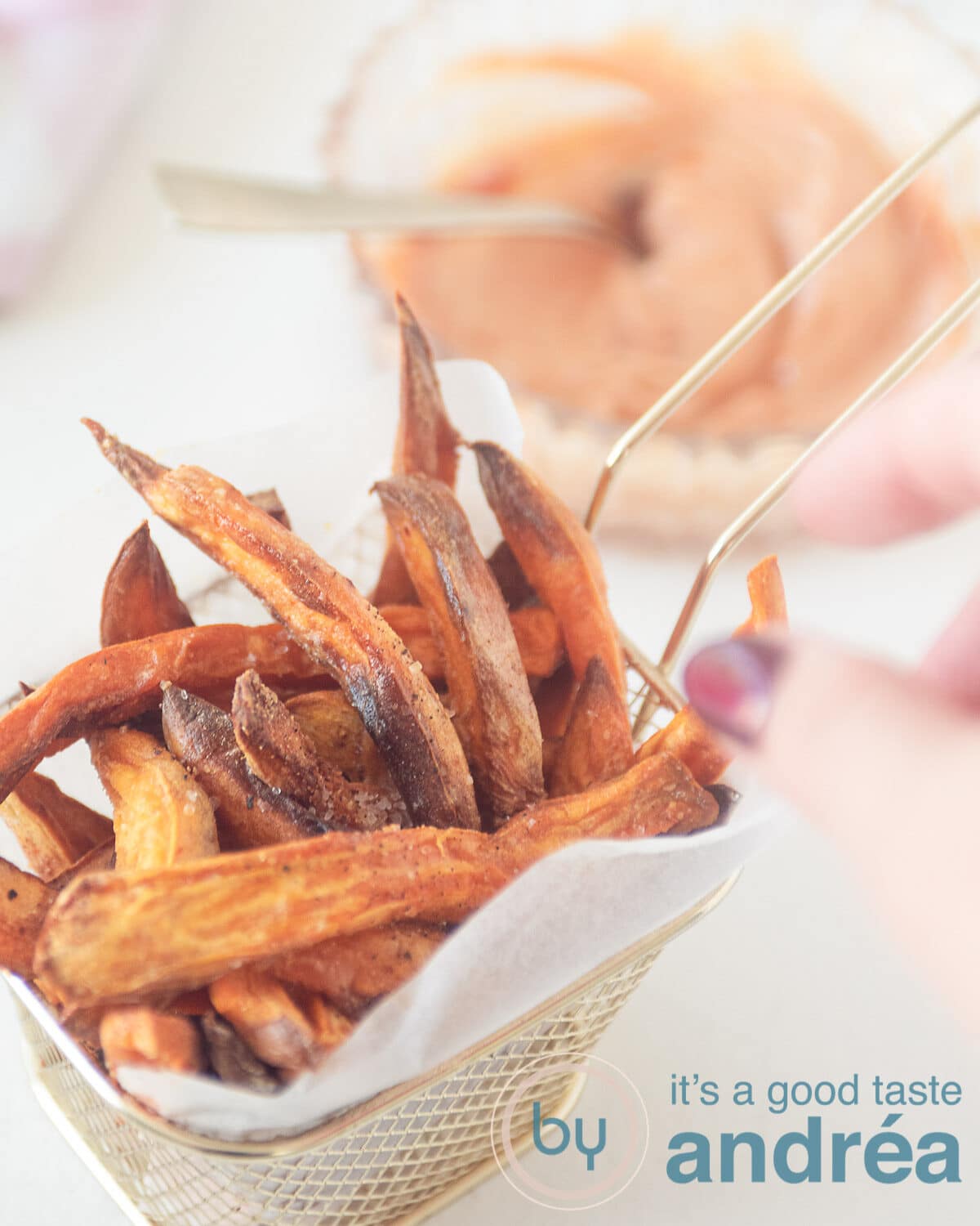 A hand taking a sweet potato fries from a golden chip tray with baking paper (which is filled with fries) in the background a dipping sauce
