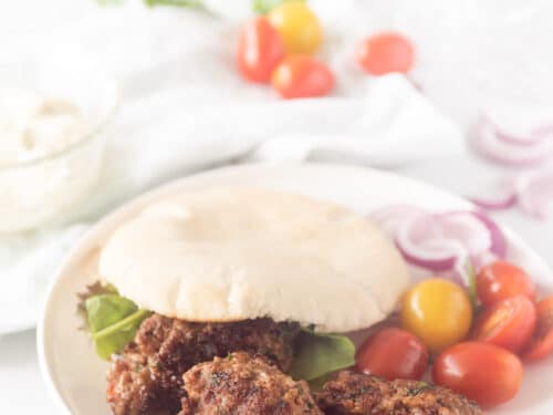 Een wit bord met een pita broodje gevuld met cevapi, sla en tomaatjes. Wat groente ligt er om het bord