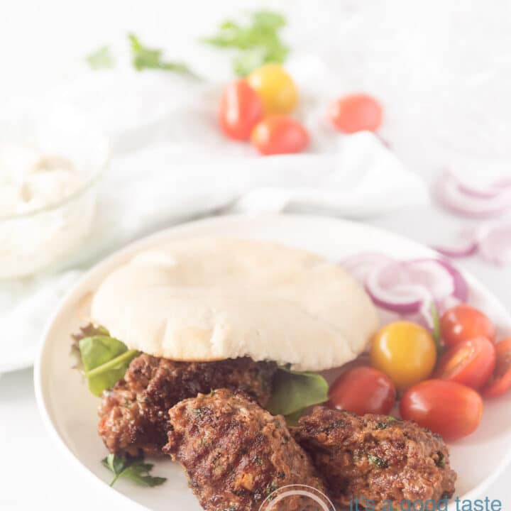 Een wit bord met een pita broodje gevuld met cevapi, sla en tomaatjes. Wat groente ligt er om het bord