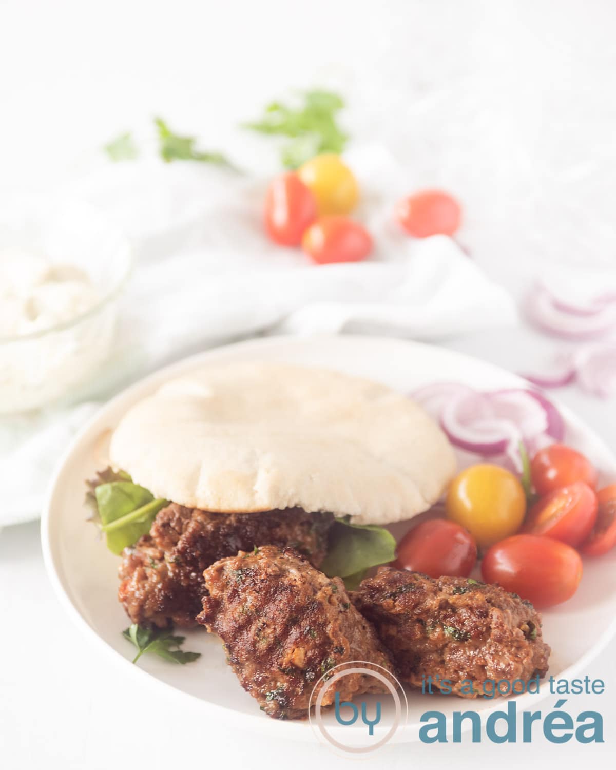 Een wit bord met een pita broodje gevuld met cevapi, sla en tomaatjes. Wat groente ligt er om het bord