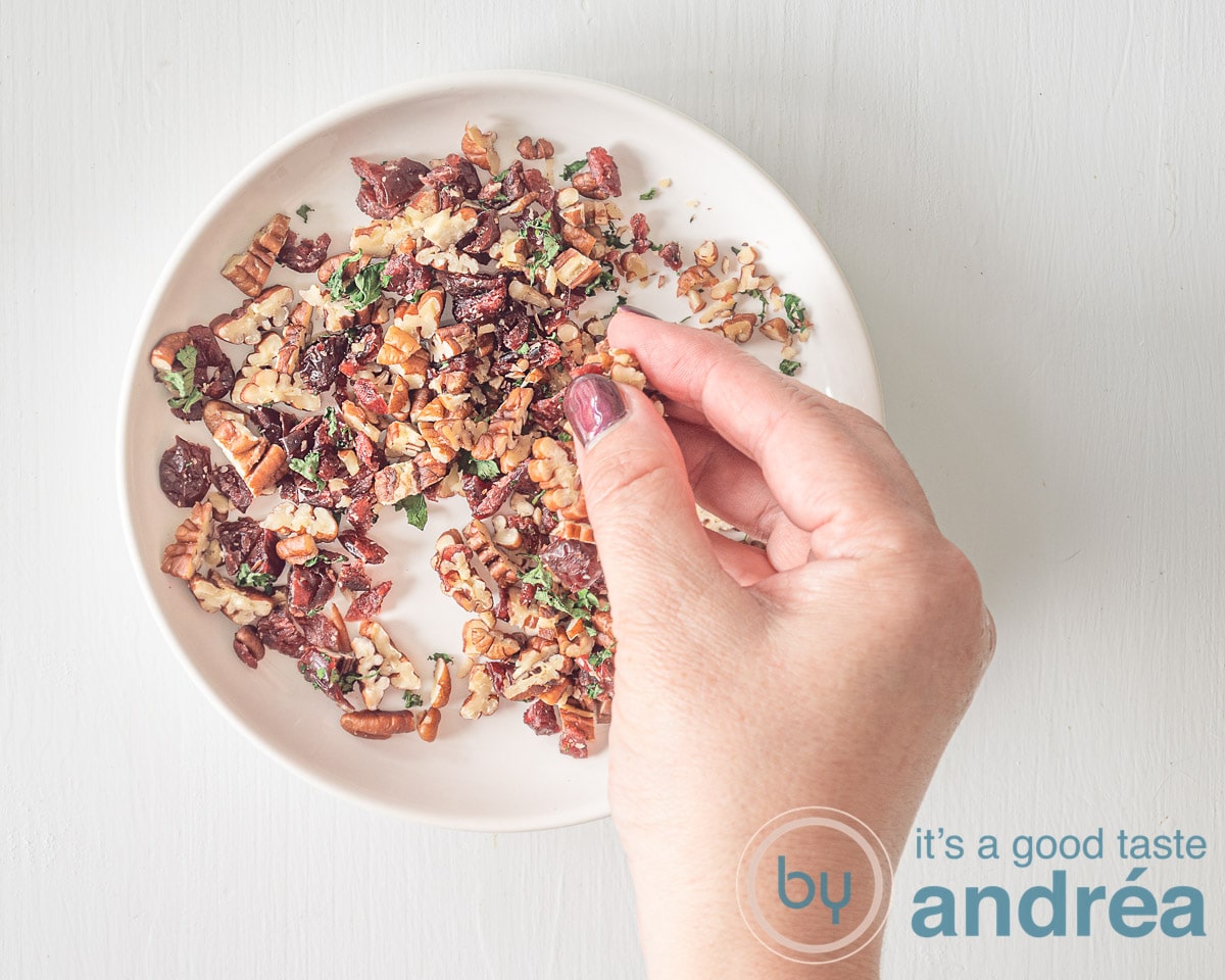 Fijngehakte cranberries, pecans en peterselie op een bord. Een hand die alles mengt.