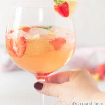 Een hand die een glas strawberry gin cocktail vast pakt met een garnering van aardbei en citroen