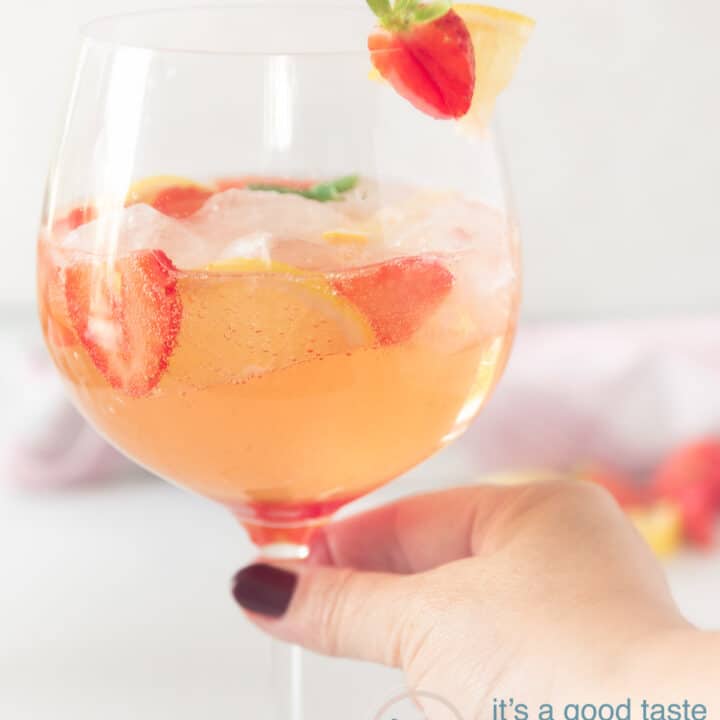 Een hand die een glas strawberry gin cocktail vast pakt met een garnering van aardbei en citroen