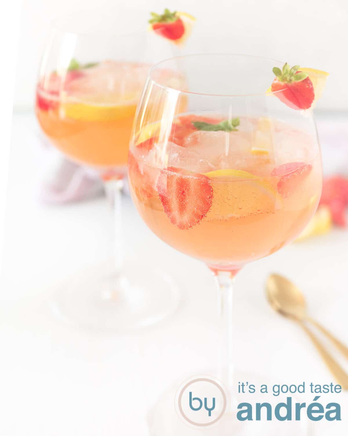 Twee glazen gevuld met een gin cocktail van aardbeien achter elkaar. Twee gouden lepels aan de zijkant.