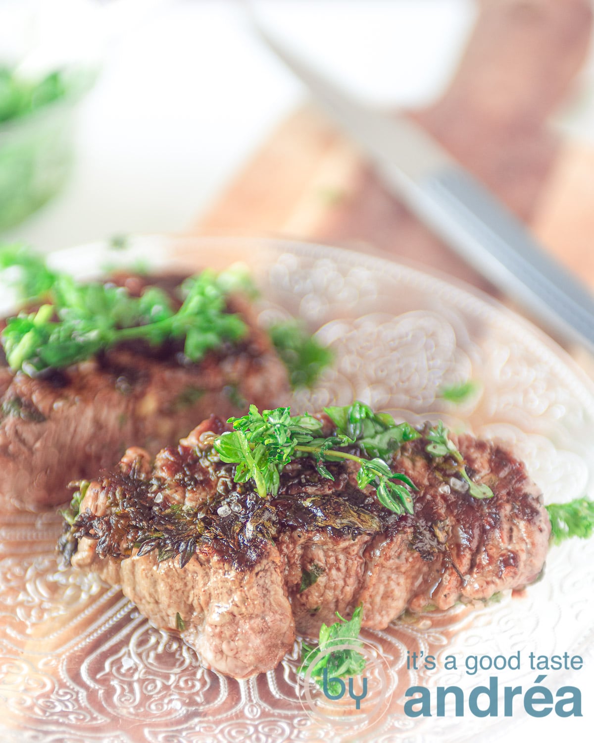 Twee gegrilde bieflapjes met groene kruiden op een glazen bord dat op een houten plank staat. Een steakmes op de achtergrond